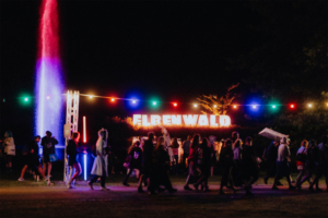 Beleuchtete Kulisse des Elbenwald Festivals bei Nacht, mit dem großen Schriftzug "ELBENWALD" im Hintergrund und bunten Lichtern, die eine lebendige Atmosphäre schaffen. Besucher in kreativen Kostümen schlendern durch das Festivalgelände.
