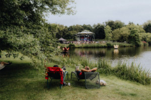 Zwei Festivalbesucher entspannen in Liegestühlen am Ufer eines Sees, während eine Bühne im Hintergrund Musik spielt. Grüne Umgebung und eine entspannte Atmosphäre prägen die Szene beim Elbenwald Festival.