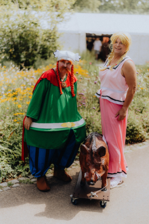 Zwei verkleidete Teilnehmer des Elbenwald Festivals stehen in einem blühenden Garten. Der linke Teilnehmer trägt ein grünes Kostüm mit rotem Umhang und einer charakteristischen Mütze, während der rechte in einem pinken Outfit posiert. Ein Holzschwein ergänzt das Bild.
