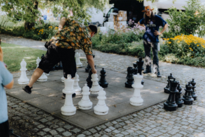 Ein Teilnehmer des Elbenwald Festivals spielt Schach mit übergroßen Figuren auf einem Außenplatz, während ein Zuschauer aufmerksam zuschaut. Die bunte Umgebung und die entspannte Atmosphäre spiegeln die kreative Vielfalt des Festivals wider.