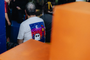 Ein Besucher des Elbenwald Festivals trägt ein T-Shirt mit einem bunten, pixeligen Gespenst-Motiv. Im Hintergrund sind weitere Festivalbesucher und bunte Sitzgelegenheiten zu sehen, die zur kreativen und lebendigen Atmosphäre des Events beitragen.