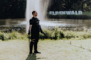 Ein Teilnehmer des Elbenwald Festivals steht am Ufer eines Teichs, gekleidet in ein schwarzes Outfit mit einem Schwert in der Hand. Im Hintergrund ist das Festival-Schild „ELBENWALD“ zu sehen, umgeben von grüner Natur und festlicher Atmosphäre.