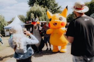 Besucher des Elbenwald Festivals posieren fröhlich in Kostümen, darunter ein überdimensionaler Pikachu und ein Drache. Im Hintergrund sind Stände und Festivalbesucher zu sehen, die die lebendige Atmosphäre des nerdigen Events genießen.