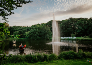 Schöne Landschaft beim Elbenwald Festival mit einem Wasserbrunnen im Vordergrund, umgeben von Bäumen und Grünflächen. Im Hintergrund ist das Festival-Schild "ELBENWALD" zu sehen, das zur einzigartigen Atmosphäre des Events beiträgt.