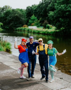 Vier verkleidete Festivalbesucher stehen am Ufer eines Gewässers. Sie tragen bunte Kostüme, inspiriert von Videospielcharakteren, und posieren fröhlich für die Kamera. Die grüne und blaue Naturkulisse des Elbenwald Festivals ergänzt die lebendige Szene.