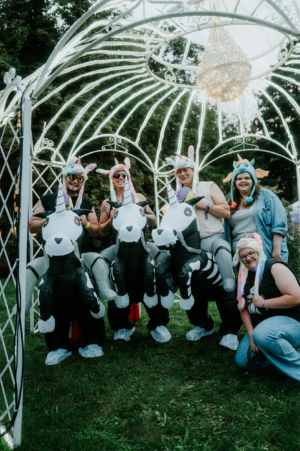 Fünf Personen in aufblasbaren Einhorn-Kostümen mit bunten Hüten posieren vor einem festlich beleuchteten Pavillon beim Elbenwald Festival. Die fröhliche Atmosphäre und die kreativen Outfits spiegeln den nerdigen Charakter des Festivals wider.