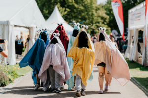 Eine Gruppe von verkleideten Festivalbesuchern in bunten Umhängen mit Augen und Tentakeln beim Elbenwald Festival. Die Teilnehmer laufen fröhlich auf dem Festivalgelände, umgeben von Zelten und anderen Besuchern.