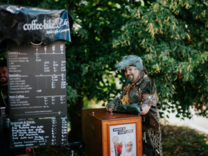Ein verkleideter Festivalbesucher mit einem krokodilähnlichen Kostüm steht lächelnd an einem Kaffee-Stand auf dem Elbenwald Festival. Im Hintergrund ist ein Menü mit verschiedenen Kaffee- und Eistee-Optionen sichtbar.
