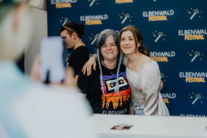 Bina Bianca Zwei Teilnehmerinnen posieren lächelnd vor dem Elbenwald Festival-Backdrop. Eine trägt ein T-Shirt mit dem Festival-Logo, während die andere in einem eleganten Outfit gekleidet ist. Im Hintergrund ist eine weitere Person zu sehen, die ein Foto macht.