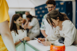Bina Bianca Eine lächelnde junge Frau interagiert mit einer Mitarbeiterin am Stand des Elbenwald Festivals, während im Hintergrund andere Besucher zu sehen sind. Die Atmosphäre ist entspannt und freundlich, ideal für Fans von Musik und Nerd-Kultur.