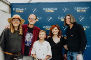 Peter Flechtner, Franciska Friede und Martin Sabel Gruppenfoto von Festivalbesuchern auf dem Elbenwald Festival, einer Mischung aus Musikfestival und Convention. Fünf Personen posieren vor einem Banner mit dem Festival-Logo, gekleidet in fantasievolle Outfits, und zeigen Freude und Gemeinschaft.