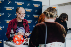 Peter Flechtner Lächelnder Mann mit Brille und rotem Hemd steht an einem Tisch beim Elbenwald Festival, während er mit einer Person spricht. Im Vordergrund sind ein D20-Würfel und eine Getränkeflasche zu sehen, die die nerdige Atmosphäre des Festivals unterstreichen.