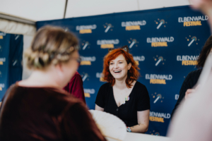 Franciska Friede Lächelnde Frau mit rotem Haar und Katzenohren, die an einem Tisch sitzt, umgeben von Festivalbesuchern. Im Hintergrund ist das Elbenwald Festival-Logo sichtbar. Die Atmosphäre ist freundlich und einladend, ideal für Fans von Musik und Nerd-Kultur.