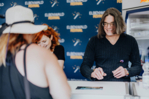 Franciska Friede und Martin Sabel Personen am Elbenwald Festival stehen an einem Stand, während sie lächeln und miteinander interagieren. Im Hintergrund ist das Festival-Logo sichtbar, das die nerdige Atmosphäre des Events unterstreicht.