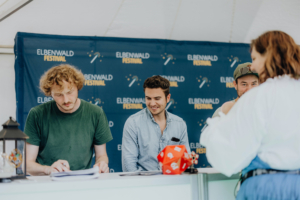 Versengold Drei junge Männer sitzen an einem Tisch auf dem Elbenwald Festival, umgeben von Festival-Dekor. Der Mann links schreibt, während die anderen lächeln und mit einer Person außerhalb des Rahmens interagieren. Ein roter Würfel liegt auf dem Tisch.