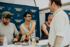 Versengold Schauspieler und Fans interagieren am Signierstand des Elbenwald Festivals, umgeben von einer bunten Festivalatmosphäre. Die Teilnehmer genießen die Mischung aus Musik und Nerdkultur, während sie Autogramme sammeln und Erinnerungen schaffen.