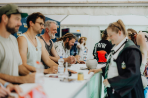 Versengold Fans und Künstler beim Elbenwald Festival, einer Mischung aus Musikfestival und Convention. Im Vordergrund signieren mehrere Personen Autogramme, während ein Fan in Kostüm mit einem roten Hut im Hintergrund zu sehen ist.