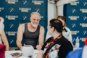 Versengold Lächelnder Mann mit grauem Bart und ärmellosem Shirt sitzt an einem Tisch beim Elbenwald Festival. Im Hintergrund ist ein Banner des Festivals zu sehen. Besucherinteraktion und freundliche Atmosphäre stehen im Fokus des Events, das Musik und Nerdkultur vereint.