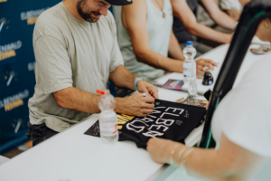 Versengold Ein Teilnehmer des Elbenwald Festivals signiert ein T-Shirt. Im Hintergrund sind weitere Personen und Wasserflaschen zu sehen. Die Atmosphäre ist entspannt und freundlich, typisch für ein kreatives Festival-Setting.