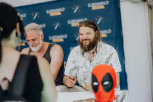 Versengold Lächelnde Teilnehmer beim Elbenwald Festival, umgeben von festlicher Atmosphäre. Im Vordergrund steht eine Deadpool-Figur, während zwei Männer am Tisch sitzen und sich mit Fans unterhalten. Das Hintergrundbanner zeigt das Logo des Festivals.