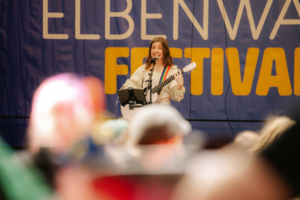 Bina Bianca Künstlerin mit Gitarre auf der Bühne beim Elbenwald Festival, umgeben von einem begeisterten Publikum. Die Veranstaltung verbindet Musik, Cosplay und die Leidenschaft für Nerd-Kultur, während die Teilnehmer die Atmosphäre genießen.