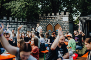 Eine Gruppe von Festivalbesuchern hebt beim Elbenwald Festival in einer lebhaften Atmosphäre die Hände. Im Hintergrund sind eine steinerne Kulisse und Biertische zu sehen, die das gemeinschaftliche und kreative Flair des Events unterstreichen.