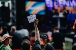 Bingo by Quizlabor Ein Festivalbesucher hebt ein Blatt Papier mit einem Plan oder einer Liste während des Elbenwald Festivals in der Menge. Im Hintergrund sind weitere Teilnehmer und eine Bühne mit bunten Lichtern zu sehen, die die lebendige Atmosphäre des Events unterstreichen.