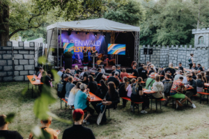 Bingo by Quizlabor Buntes Treiben beim Elbenwald Festival: Eine lebhafte Menge versammelt sich an Tischen vor der Bühne, während Künstler auftritt. Im Hintergrund sind eine aufwendige Kulisse und festliche Beleuchtung zu sehen, die die Atmosphäre des Festivals unterstreichen.