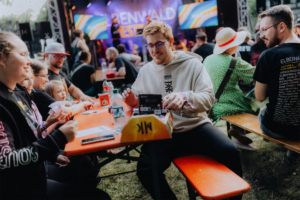 Bingo by Quizlabor Festivalbesucher beim Elbenwald Festival, die an einem orangefarbenen Tisch sitzen und an einem Spiel oder einer Aktivität teilnehmen. Im Hintergrund sind Bühnenperformances und andere Teilnehmer in nerdigen Outfits zu sehen.