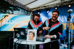 Bingo by Quizlabor Zwei Männer in bunten Anzügen stehen an einem Tisch mit Mikrofonen und einem Mischpult auf dem Elbenwald Festival. Im Hintergrund ist ein Bildschirm mit der Aufschrift "Backstreet Boys" zu sehen, der die festliche Atmosphäre des Events unterstreicht.