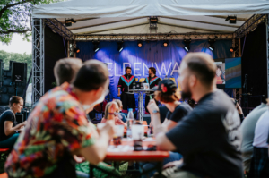Bingo by Quizlabor Zwei Moderatoren stehen auf der Bühne des Elbenwald Festivals und präsentieren vor einem Publikum, das an Tischen sitzt. Die Atmosphäre ist lebhaft, mit bunten Outfits und einer Mischung aus Musik und Gesprächen im Hintergrund.
