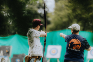 Bogenweide Ein junger Mann mit einer Bogen in der Hand zielt auf ein Ziel, während ein Trainer ihm Anweisungen gibt. Im Hintergrund sind mehrere Zielscheiben und eine grüne Kulisse zu sehen. Die Szene fängt die sportliche Atmosphäre des Elbenwald Festivals ein.