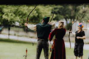 Bogenweide Teilnehmer des Elbenwald Festivals üben das Bogenschießen in fantasievoller Kleidung. Im Vordergrund sind drei Personen zu sehen, die mit Bögen zielen, umgeben von einer grünen Wiese und Bäumen im Hintergrund. Ein kreatives und interaktives Erlebnis.
