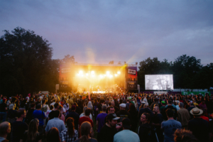 Blick auf die Bühne des Elbenwald Festivals, umgeben von einer großen Menge begeisterter Fans. Die Atmosphäre ist lebhaft, mit bunten Lichtern und einem großen Bildschirm, der eine Live-Performance zeigt, während der Himmel in sanften Farben leuchtet.