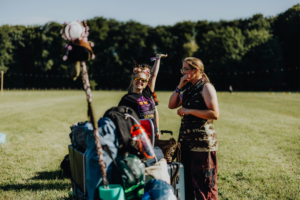 Zwei Festivalbesucherinnen stehen auf dem Elbenwald Festival-Gelände, umgeben von Gepäck. Eine trägt eine Sonnenbrille und ein T-Shirt mit Festival-Logo, während die andere nachdenklich wirkt. Im Hintergrund ist eine grüne Wiese und Bäume sichtbar.