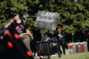 Eine Teilnehmerin des Elbenwald Festivals zieht einen Wagen mit Gepäck und einem bunten Schild mit der Aufschrift "CAMP Kunterbunt". Im Hintergrund sind andere Festivalbesucher und Toiletten zu sehen, die die lebendige Atmosphäre des Events unterstreichen.