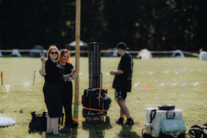 Drei Festivalbesucher am Elbenwald Festival stehen auf einer Wiese. Eine Frau mit Sonnenbrille macht das Peace-Zeichen, während eine andere lächelt. Im Hintergrund sind Zelte und bunte Wimpel zu sehen, die die lebendige Atmosphäre des Events unterstreichen.