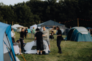 Zeltaufbau auf dem Elbenwald Festival: Eine Gruppe von Festivalbesuchern bereitet ihre Zelte vor, umgeben von einem bunten Zeltlager. Grüne Wiese und festliche Lichter im Hintergrund schaffen eine einladende Atmosphäre für das Event.