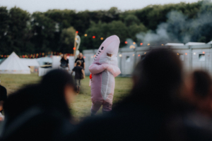 Ein Besucher im pinken Haifischkostüm steht auf dem Elbenwald Festival, umgeben von anderen Festivalbesuchern und festlicher Beleuchtung. Im Hintergrund sind Zelte und Rauch zu sehen, was die lebendige Atmosphäre des Events unterstreicht.
