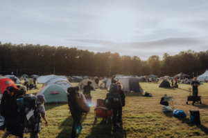 Zeltplatz beim Elbenwald Festival mit Teilnehmern, die ihre Ausrüstung transportieren. Die Szene zeigt zahlreiche Zelte unter einem klaren Himmel, umgeben von Bäumen, und vermittelt die lebendige Atmosphäre des Festivals.