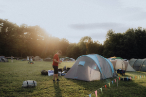 Zeltplatz beim Elbenwald Festival, mit Menschen, die sich auf das Event vorbereiten. Im Hintergrund sind zahlreiche Zelte und eine grüne Wiese zu sehen, während die Abendsonne eine gemütliche Atmosphäre schafft. Ideal für Fans von Musik und Nerdkultur.
