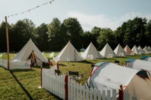 Zeltplatz des Elbenwald Festivals mit weißen Tipis und grünen Zelten, umgeben von Wiese und Bäumen. Einige Sitzmöglichkeiten und dekorative Lichter sorgen für eine einladende Atmosphäre für Festivalbesucher und Nerds.