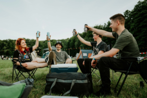 Vier Freunde heben ihre Getränke in die Luft, während sie auf Campingstühlen sitzen. Im Hintergrund sind Zelte und eine grüne Wiese zu sehen, die die entspannte Atmosphäre des Elbenwald Festivals widerspiegeln.