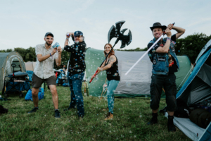 Vier Festivalbesucher auf dem Elbenwald Festival posieren spielerisch mit verschiedenen Requisiten, darunter eine Lichtschwert, eine Axt und Seifenblasen, umgeben von Zelten auf einer grünen Wiese. Die Szene strahlt eine fröhliche und kreative Atmosphäre aus.