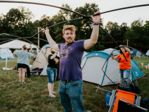 Ein junger Mann in einem lila T-Shirt hält Zeltstangen hoch, während im Hintergrund andere Festivalbesucher Zelte aufbauen. Die Szene spielt auf dem Elbenwald Festival, wo Musik und Nerdkultur aufeinandertreffen.