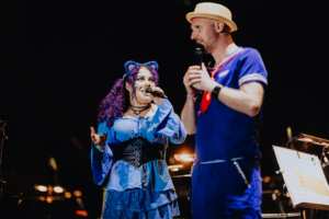SaKiDe & Olaf Unzel Bunt gekleidete Künstler auf der Bühne des Elbenwald Festivals, während sie ein Publikum mit Musik und Unterhaltung begeistern. Die Sängerin trägt ein auffälliges blaues Outfit und hat lila Haare, während der Moderator in einem maritimen Look auftritt.