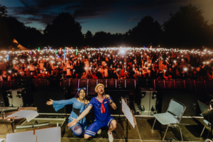 SaKiDe & Olaf Unzel Zwei Performer auf der Bühne des Elbenwald Festivals, umgeben von einer begeisterten Menge, die mit ihren Handys leuchtet. Die Szene zeigt die energiegeladene Atmosphäre des Festivals, das Musik und Nerd-Kultur verbindet.