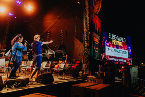 EWF 2026! Bühne beim Elbenwald Festival mit zwei Performern, die das Publikum ansprechen. Im Hintergrund ist ein großes Banner mit den Festival-Daten vom 7. bis 9. August 2026 im Spreeauenpark Cottbus zu sehen. Stimmungsvoll beleuchtet, voller Vorfreude.