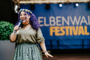SaKiDe Eine fröhliche Moderatorin mit lila Haaren und Blumenkranz hält ein Mikrofon in der Hand, während sie auf der Bühne des Elbenwald Festivals spricht. Im Hintergrund ist das Festival-Logo gut sichtbar. Die Atmosphäre ist lebhaft und einladend.