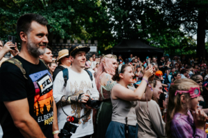 Eine begeisterte Menge von Festivalbesuchern beim Elbenwald Festival, das eine Mischung aus Musik und Nerdkultur bietet. Die Teilnehmer, in bunten Outfits, filmen und fotografieren die Auftritte auf der Bühne, um die besondere Atmosphäre festzuhalten.